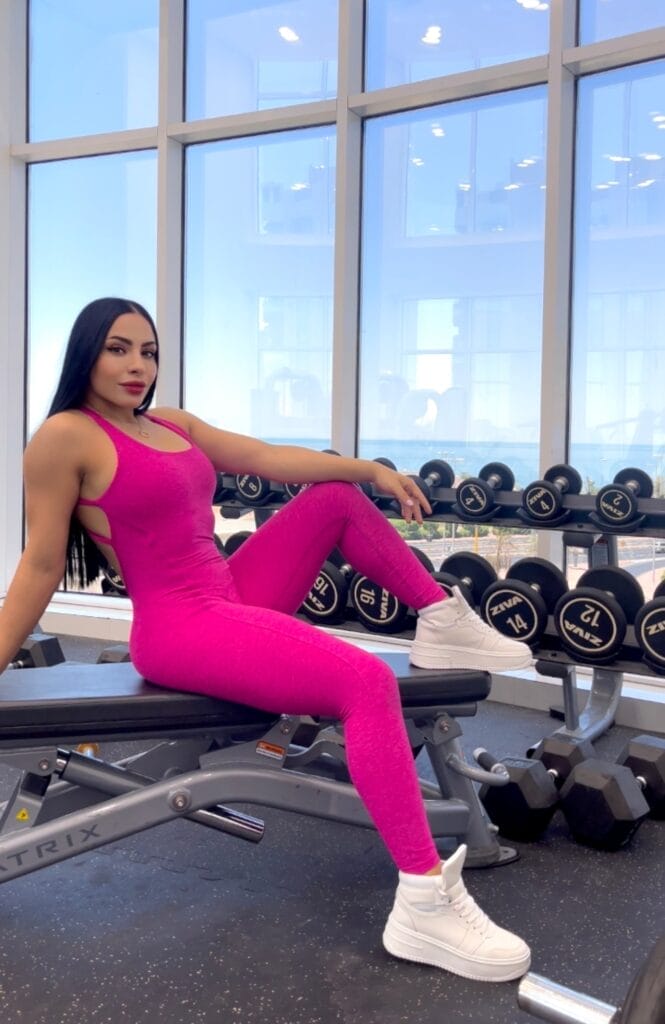 A person in a bright pink athletic outfit sits confidently on a gym bench, surrounded by dumbbells. The gym has large windows, allowing natural light to stream in. They are wearing white sneakers and have long dark hair, with a composed expression.
