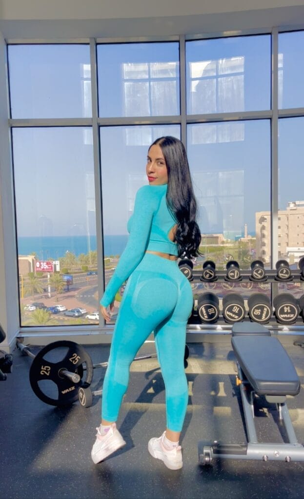 A person with long dark hair, wearing a bright blue workout set and white sneakers, stands in a gym. They face backward, looking over their shoulder. Behind them, there are large windows with a city and ocean view. Gym equipment, such as dumbbells and a bench, is visible.