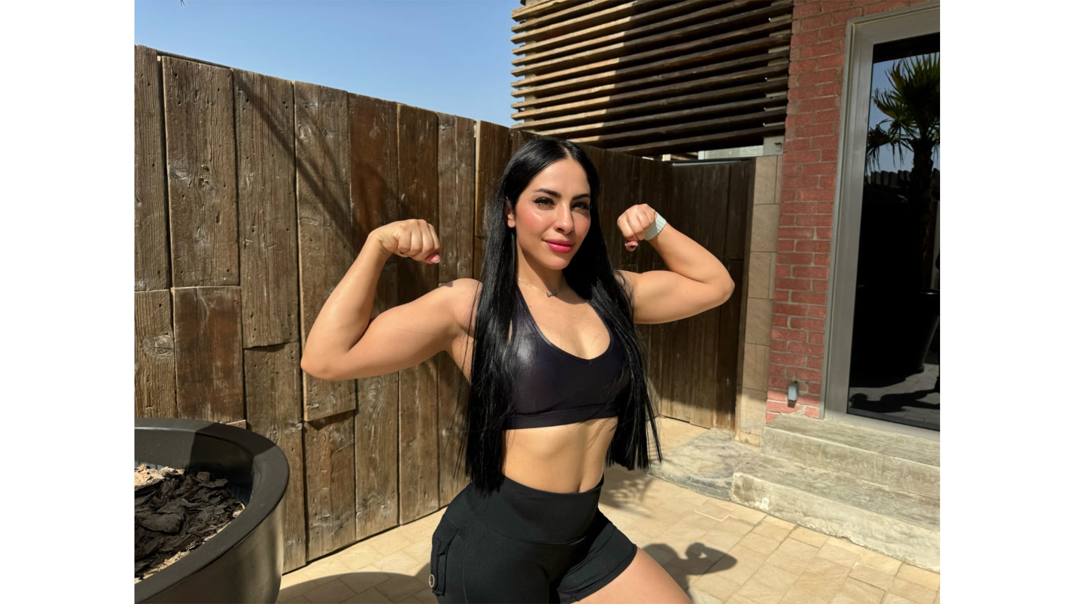 A woman with long black hair poses outdoors, flexing her arms to show her muscles. She wears a black sports bra and black shorts. The wooden fence and modern building in the background suggest a sunny day in a courtyard or patio area. Shadows indicate bright lighting conditions.